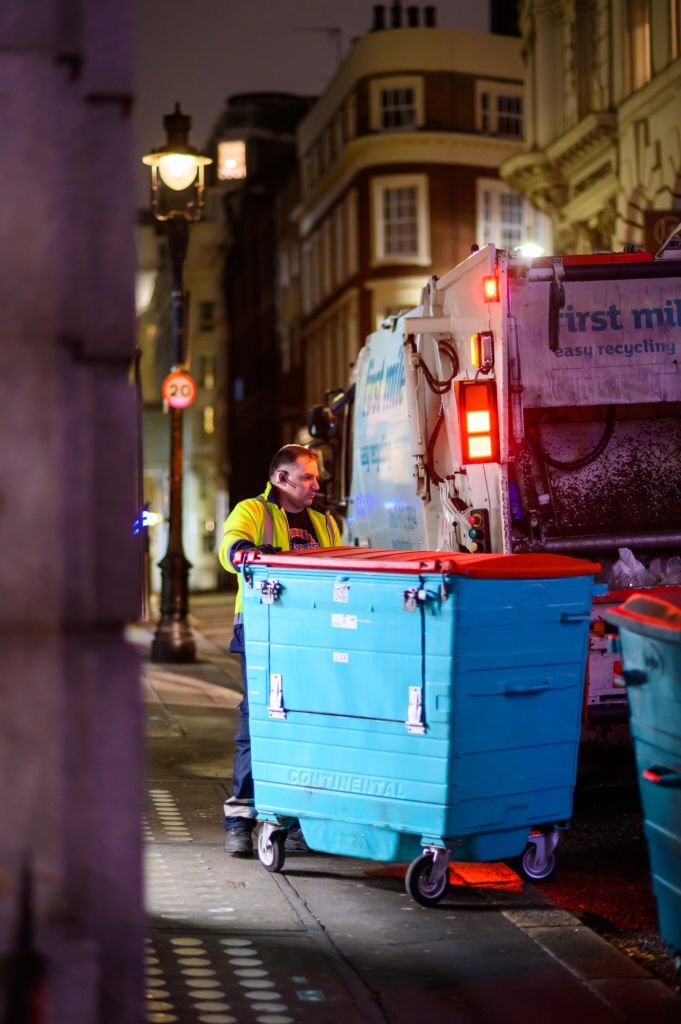 Bin man pushes large wheelie bin at back of bin lorry at night